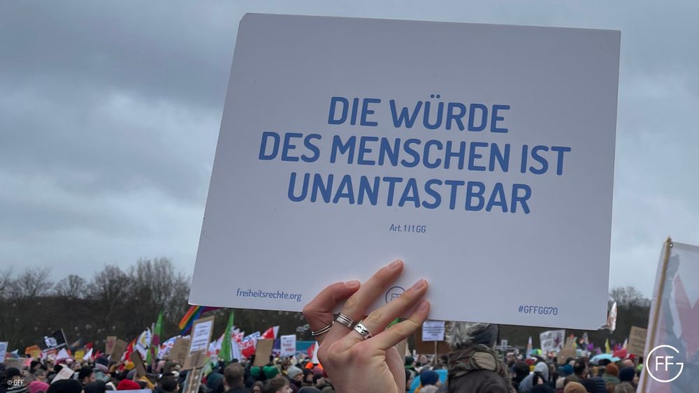 Ein Schild mit der Aufschrift „Die Würde des Menschen ist unantastbar, Art. 1 I 1 GG“ in die Höhe gehalten – im Hintergrund eine Menschenmenge.