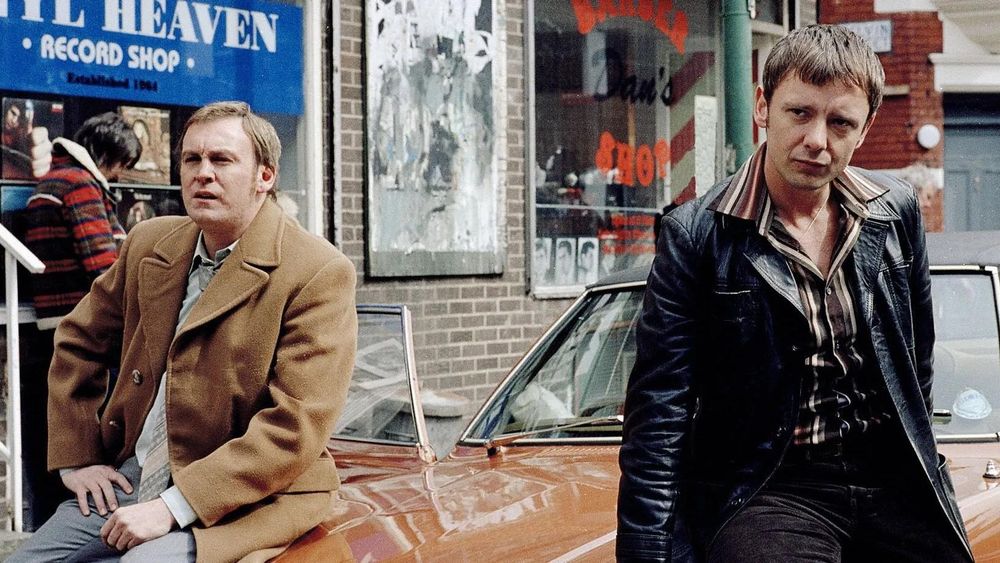Life on Mars actors John Simm and Philip Glenister sitting on a brown Ford Cortina outside of a record shop in (supposedly) early 1970s Manchester, England.