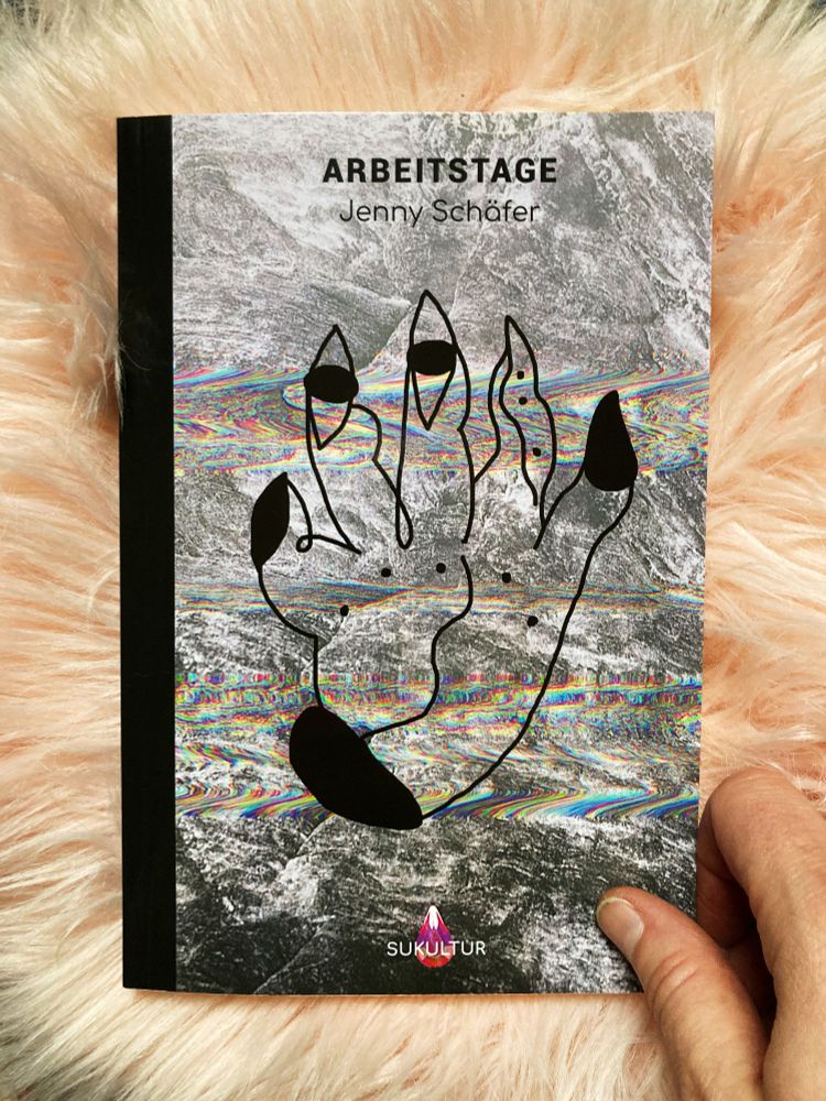 Das Buch Arbeitstage von Jenny Schäfer liegt auf einem Fellteppich und wird von einer Hand gehalten. Das Buchcover zeigt einen Glitch auf einer Gesteinsfotografie, einen Glitzerstein und eine surreale Handzeichnung.