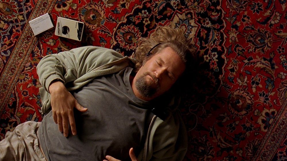 Overhead shot of Jeff Bridges lying down on an Oriental rug from the movie ‘The Big Lebowski’. His eyes are closed while listening via headphones to a Walkman that lies to his right side. A cassette case is also next to the Walkman. He’s wearing grey sweats.