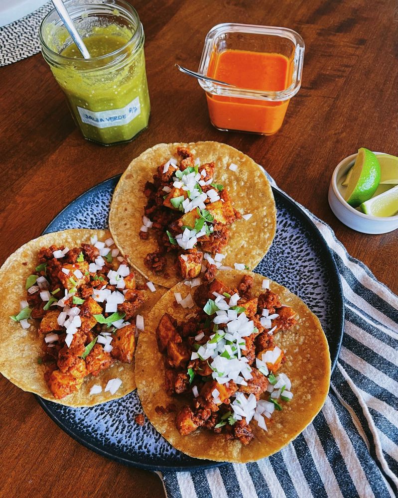 Three tacos on a plate on a table filled with vegan chorizo and potato topped with white onion and cilantro surrounded by containers of salsa verde, salsa roja, and limes. 