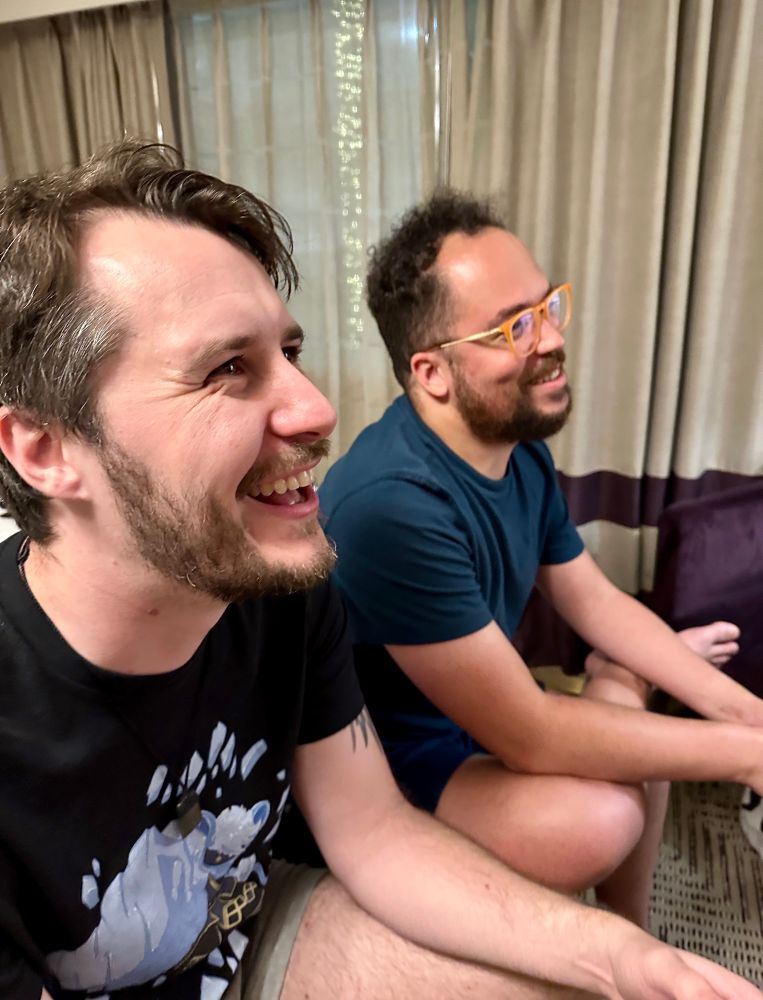 two men holding video game controllers and staring out of frame to the right, they are smiling and having fun now