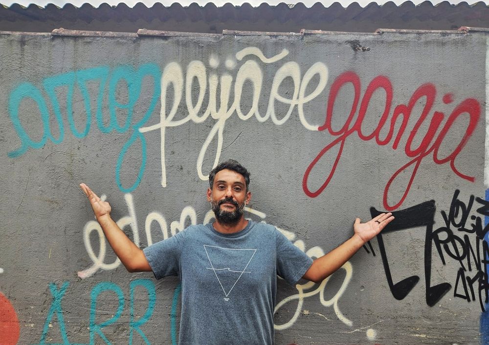 Eu com os braços abertos encostado em um muro, e logo acima de mim o grafite com os dizerez "arroz feijão e ganja"