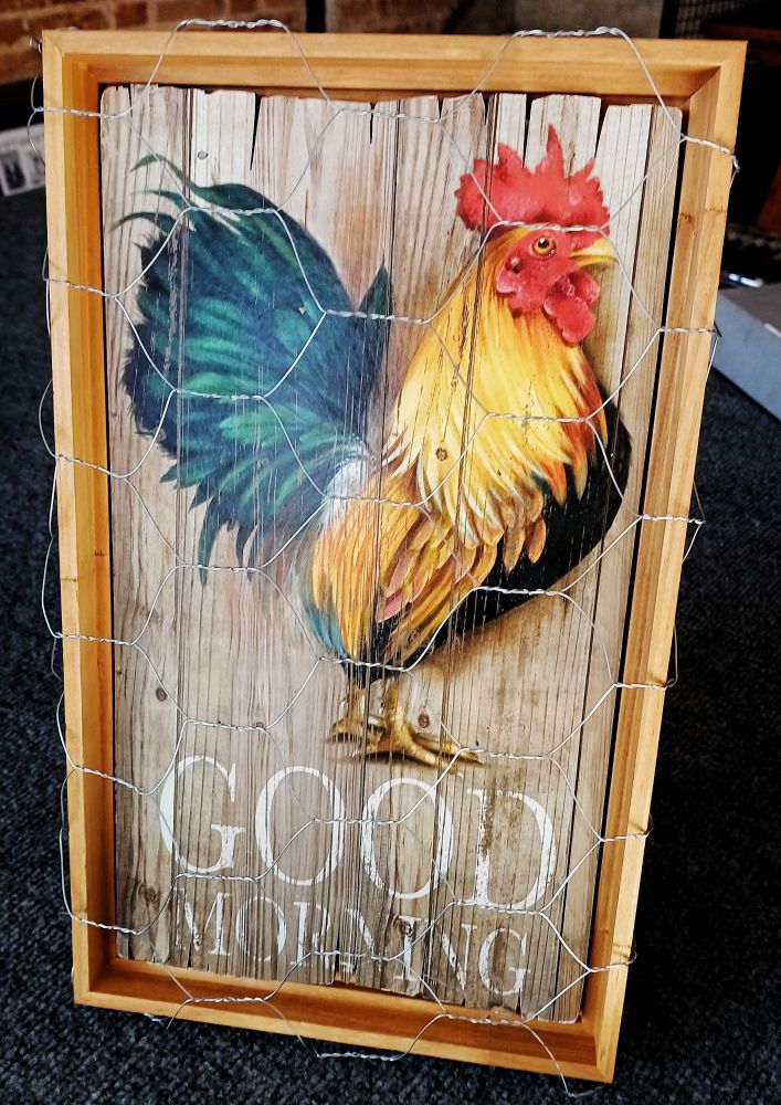 Uma placa com desenho de um galo e as palavras Good Morning, dentro de uma moldura com cor de madeira, com uma tela de arame na frente.