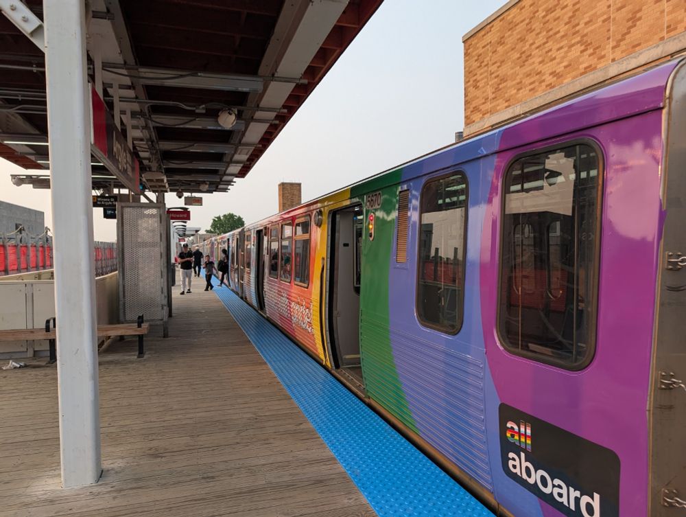 A picture of the 2025 CTA Pride Train with bold, wavy stripes of color, again aligned to put the Black and brown stripes and trans stripes at the front end of each train by ensuring they appear at the cab ends of each car and thus are forward no matter which way the train is rolling along the tracks, with the classic pride colors right in step behind them.