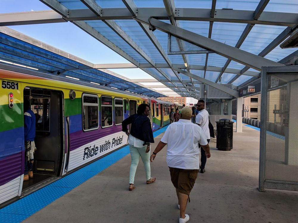The 2018 Pride Train with a wavy pattern of the classic six pride colors and the words "Ride with pride!" below the window line as people exit the train at the Wilson station. Bonus detail: my partner and family are in the foreground since we were taking the Red Line together!