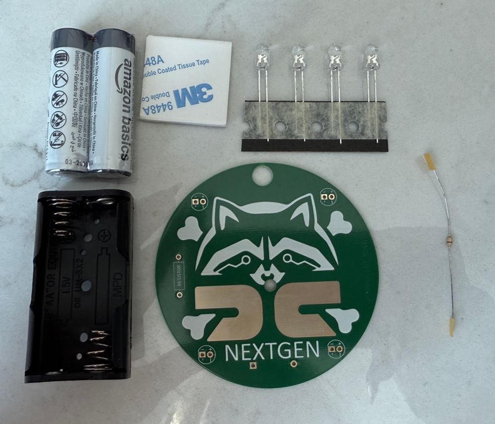 Overhead view of an electronics kit for @defconnextgen laid out on a white surface. The kit includes two Amazon Basics AA batteries, a square of 3M 9448A double-coated tissue tape, four clear LEDs attached to a strip of paper, a black plastic AA battery holder, a circular green printed circuit board (PCB) with a raccoon face and the text ‘DC NEXTGEN’ in gold, and a single resistor with yellow tape on each lead.