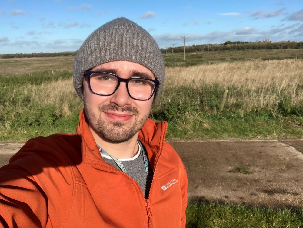 A young man wearing glasses and a warm hat, with the open flat landscape of the Cambridgeshire Fens behind him.  A wide expanse of grassland and agricultural land.