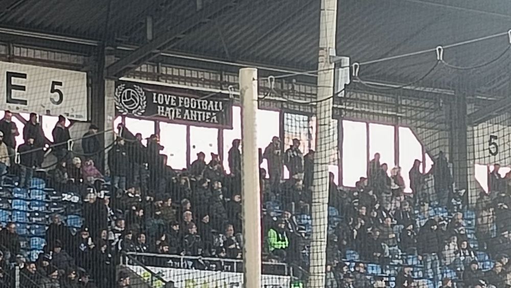 Foto. Blick auf Block E5 im Stadion von Waldhof Mannheim, viele Waldhof-Fans. Ganz oben hänt ein Banner mit weißer Schrift auf schwarzem Grund: "LOVE FOOTBALL, HATE ANTIFA".