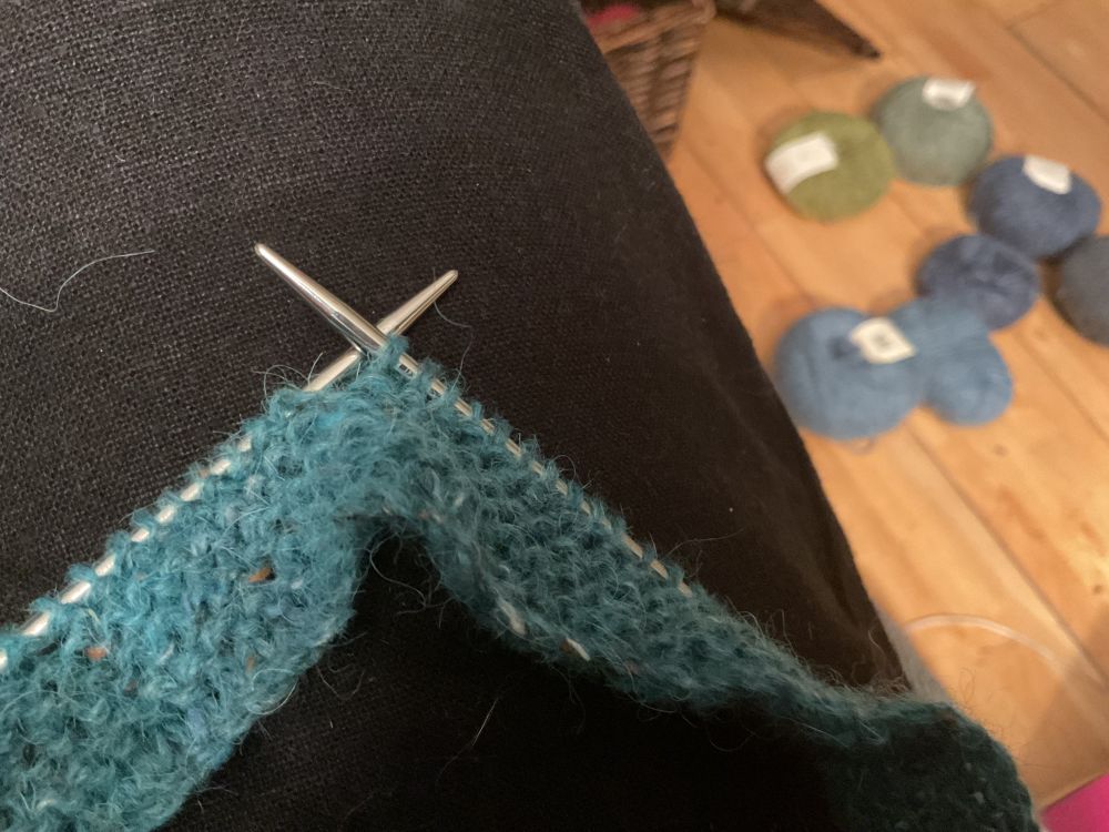 A few rows of blue-green yarn on silver needles, other blue and green balls of yarn can be seen in the background