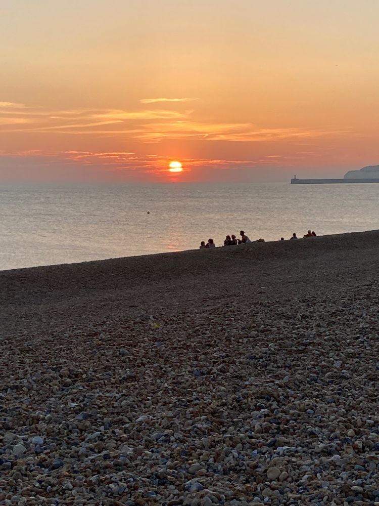 Sunset from the pebble beach in Seaford, UK. It’s nearly dark, but the outlines of a small group of friends are silhouetted against the grey, silver sea. 