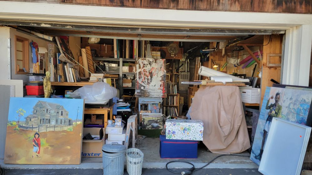 An open two-car garage with no cars in it. Easel, table, printmaking materials, garbage, donations, and a couple visible paintings in it.