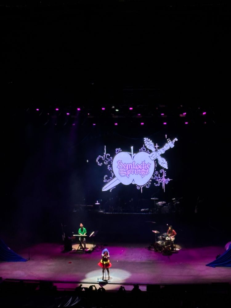 a photo of a stage lit up pink with 3 people on stage and a logo showing an apple with a sword through it that says hemlocke springs 