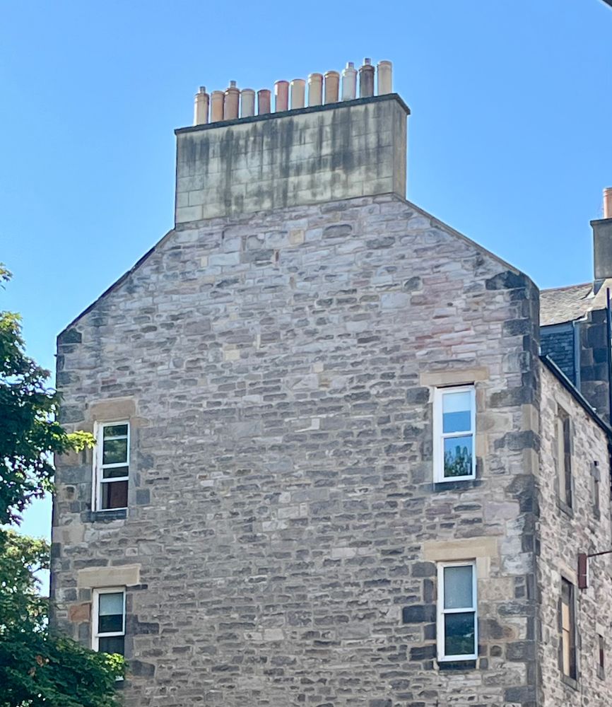 View of the windows at the end of a building in Edinburgh old town, with a part of a tree to the left. The stones of the building are various shades of brown. Chimney-pots can be seen on the roof. 