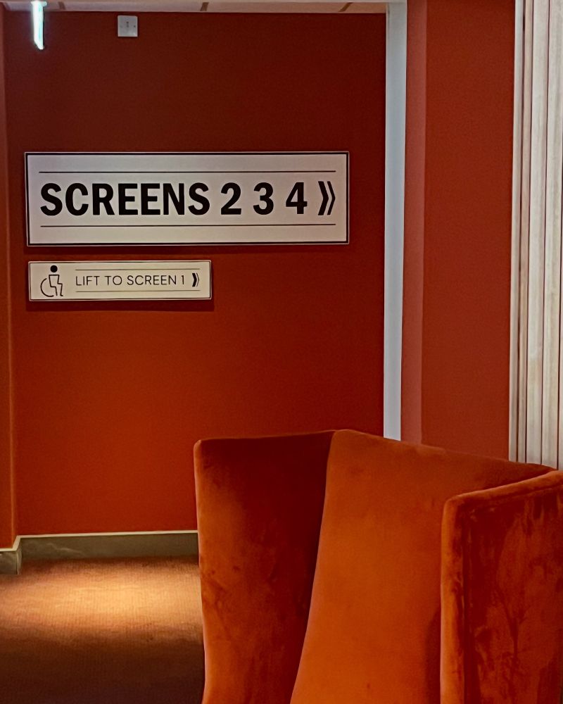 In the foreground, a red velvet chair, behind which is a passageway with a sign directing people to screens 2,3 and 4 and the lift to screen 1. The walls and carpet are in the same red as the chair. 