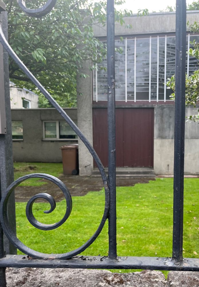  View through the curlicues on a wrought iron railing towards the large brown doors of a late 20the century church building. Trees, lawn and a brown garden waste bin are also visible. 