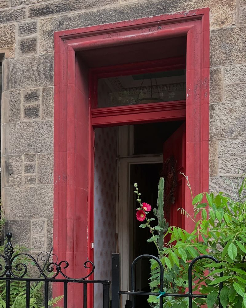 An open red door with a red surround, leading into a small garden with lots of greenery and red hollyhocks in bloom near the centre of the picture. 