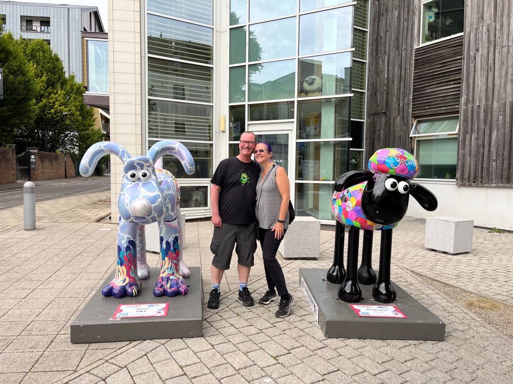 Ocean Gromit covered in sea shells on the left. Paul & Dawn in the middle with big grins. Shaun the sheep with bright colors on the right. Aardman studios in the background. 