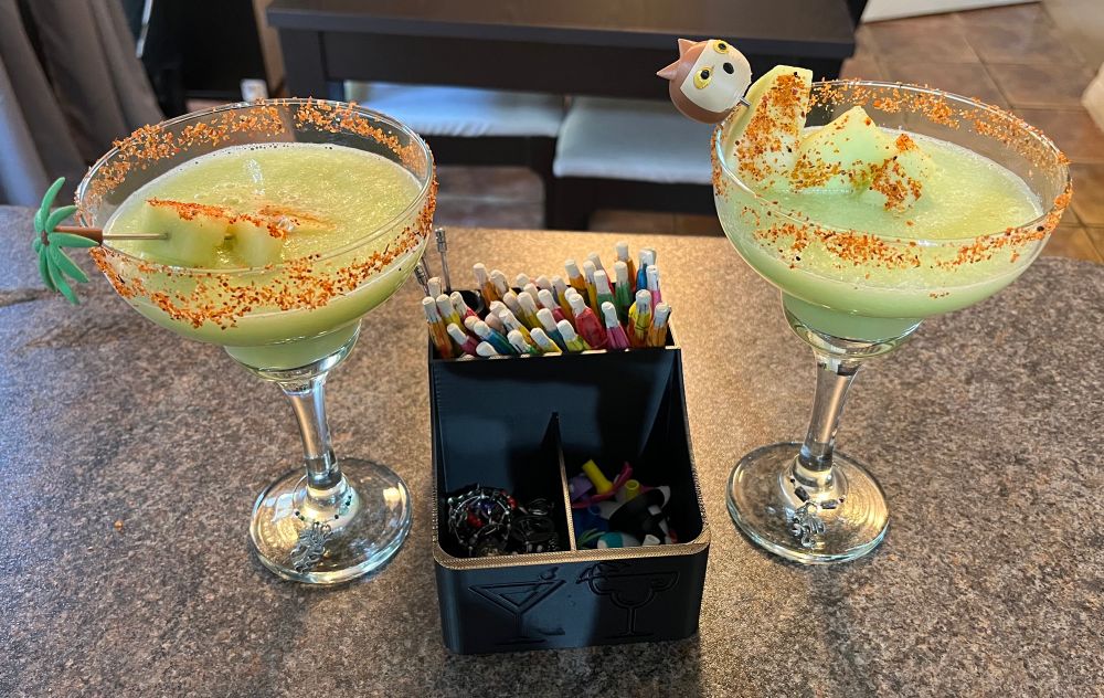 2 margaritas on either side of a drink accessory box wine charms, cocktail sticks, and 3D printed stick toppers. The left margarita has a palm tree topper and unicorn wine charm. The right has an owl bear topper and dragon charm. 