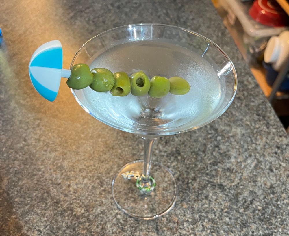 Martini with 6 nocellara olives on a metal stick topped with a blue and white beach umbrella and a gecko wine charm on the stem. 