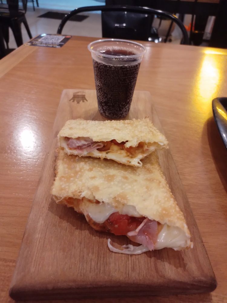 Foto de um pastel cortado ao meio, mostrando queijo, tomate e presunto, e um copo de coca atrás