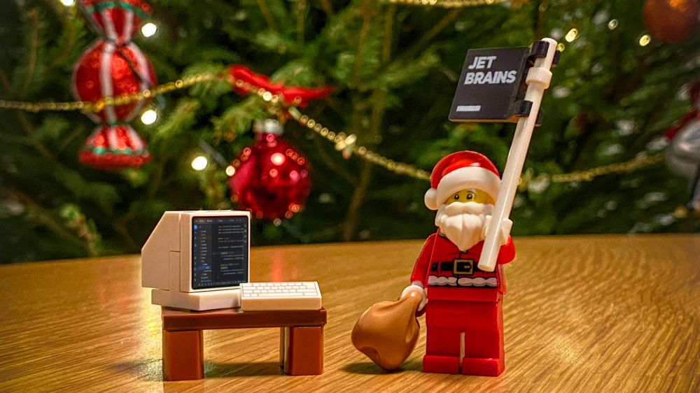 A festive scene featuring a LEGO Santa Claus figurine holding a "JetBrains" flag and a sack. Next to Santa is a miniature LEGO-style computer on a small wooden table, with a monitor displaying code in a JetBrains IDE. The background includes a decorated Christmas tree adorned with red ornaments and glowing lights.