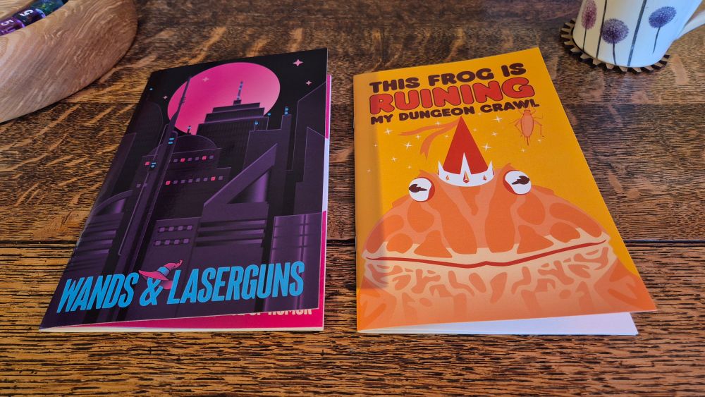 Front covers of two zines. Left: Wands & Laserguns, featuring a futuristic neon city. Right: This Frog is Ruining my Dungeon Crawl, featuring a large frog with a small crown
