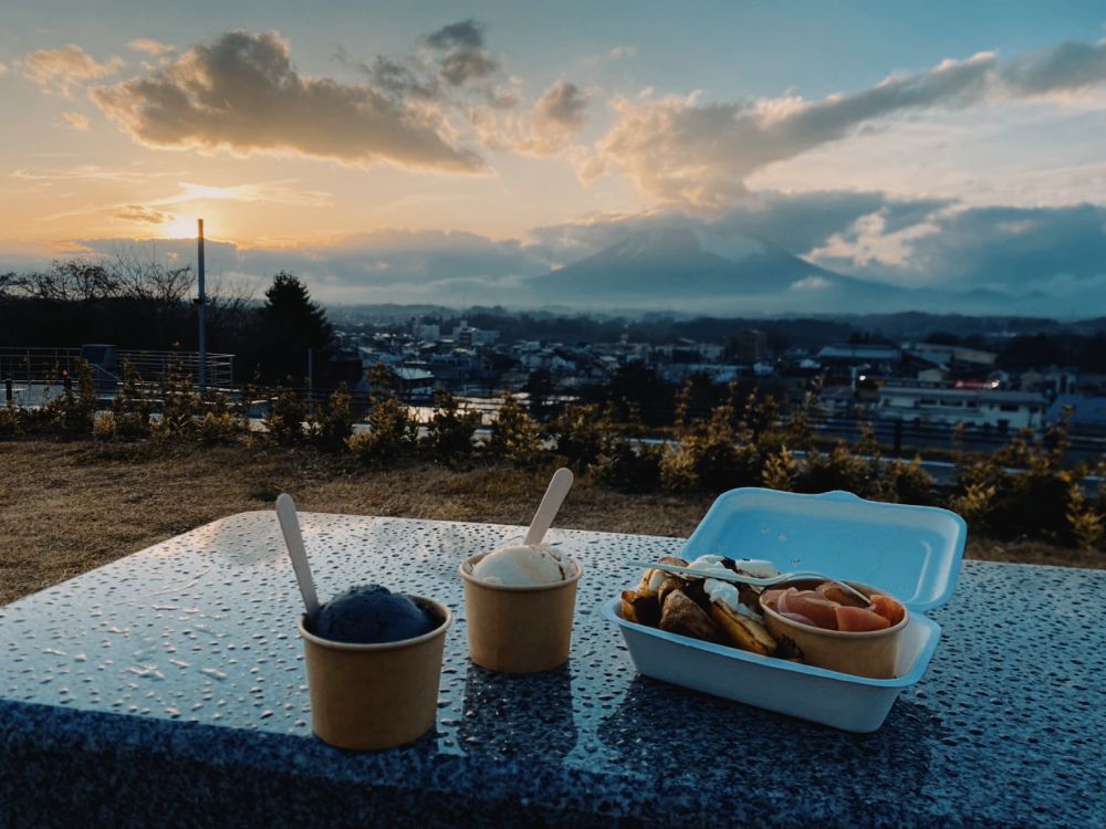 見晴らしのいい丘の向こうに夕暮れ時の空と雲がかった岩手山の景色。手前のベンチにはアイスとプーティン（カナダの食べ物）が置かれている。