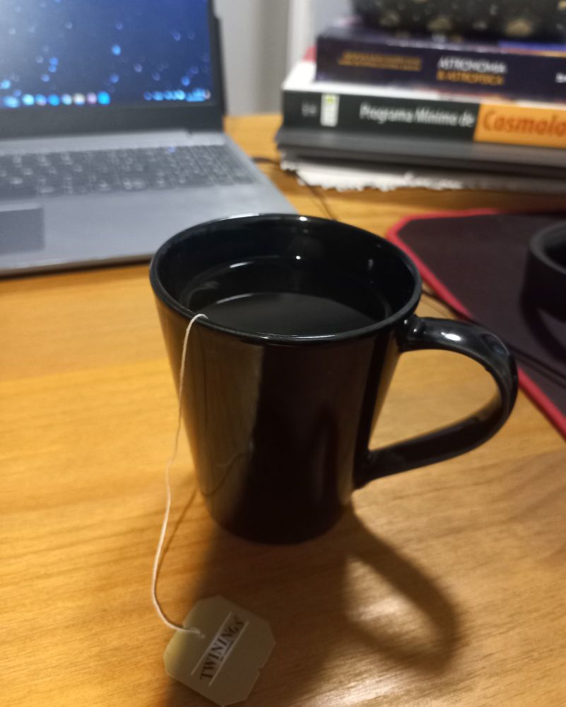 A black mug filled with Earl Grey Tea stands on a wooden table, an open laptop with showing a starry wallpaper on the background with cosmology and astronomy books by it's side.