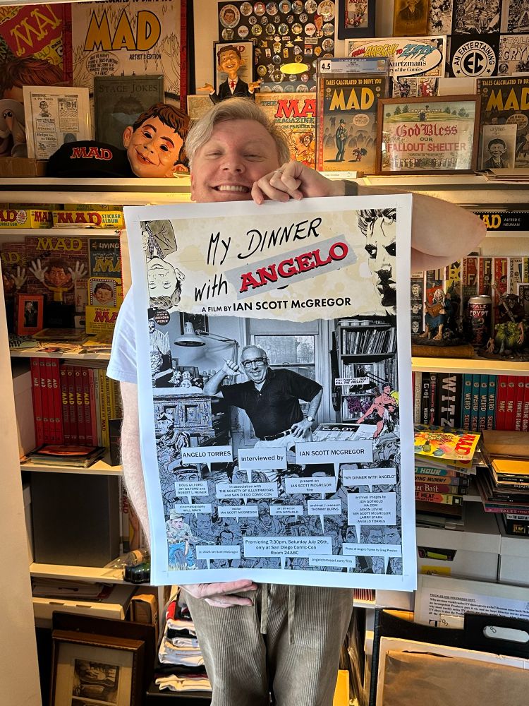 A young-at-heart aging, but attractive enough, man stands in front of a colorful collection of MAD Magazine ephemera. He smiles big and holds a movie poster for a film he directed, a documentary about comics artist Angelo Torres, called "MY DINNER WITH ANGELO"