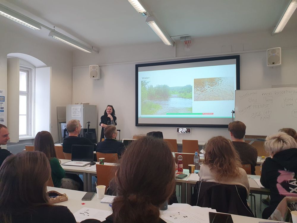 Daria Zhornik (LMU Munich) giving a presentation in front of students. The current slide shows bodies of water and has the headline "water".