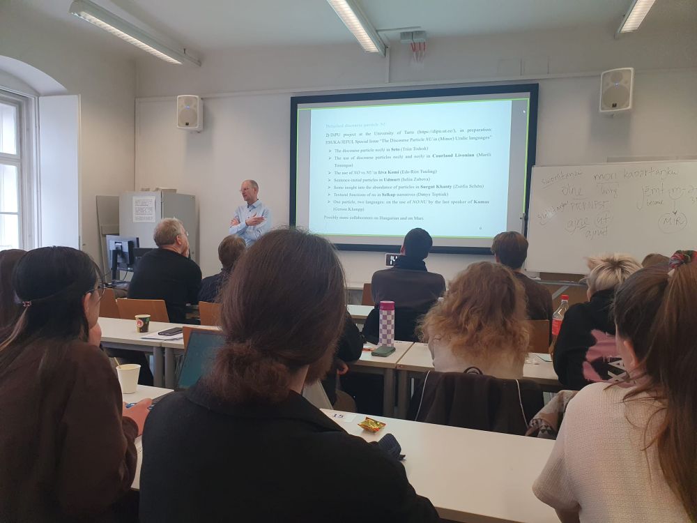 Gerson Klumpp (University of Tartu) giving a talk about discourse particles in Uralic languages in front of students. The current slide has the headline "Detached discourse particle NU" and presents the DiPU project (https://dipu.ut.ee/).