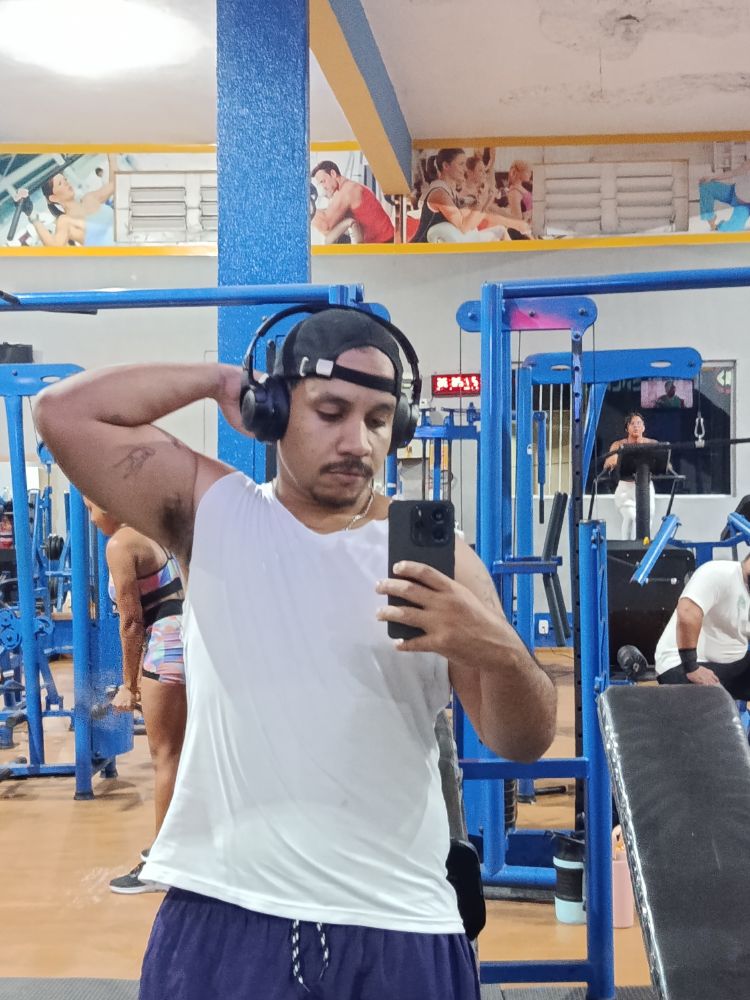 Selfie minha (homem negro) na academia. Uso camisa branca recortada, boné para trás e bermuda roxa. Estou com o braço esquerdo levantado, mostrando bíceps 
