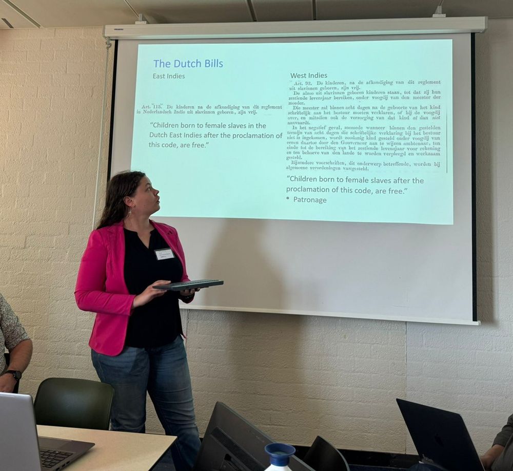 Ida stands in front of her PowerPoint presentation, which is currently on a slide about Dutch bills in the East Indies. She is wearing a pink jacket and a white conference nametag, and she looks up at the slide while she talks.