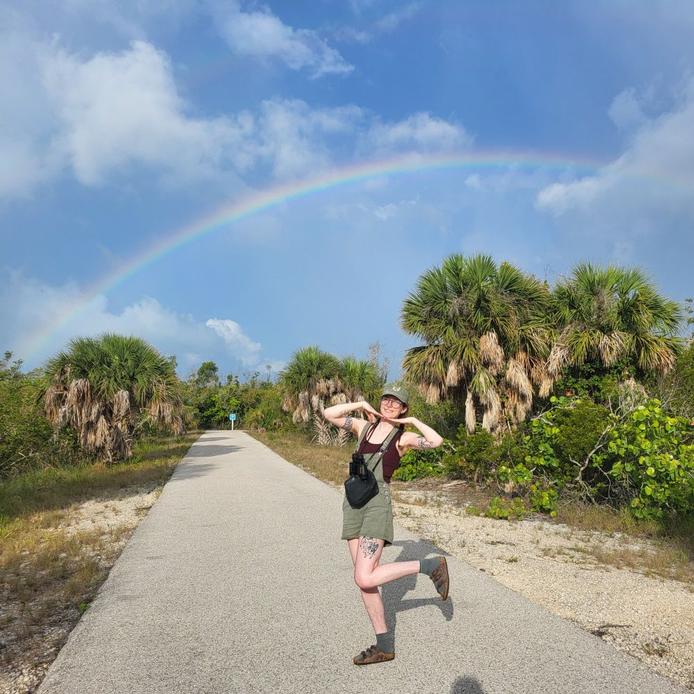 Me, a young woman in green overall shorts and a green hat, posing cutely in front of a bright rainbow 