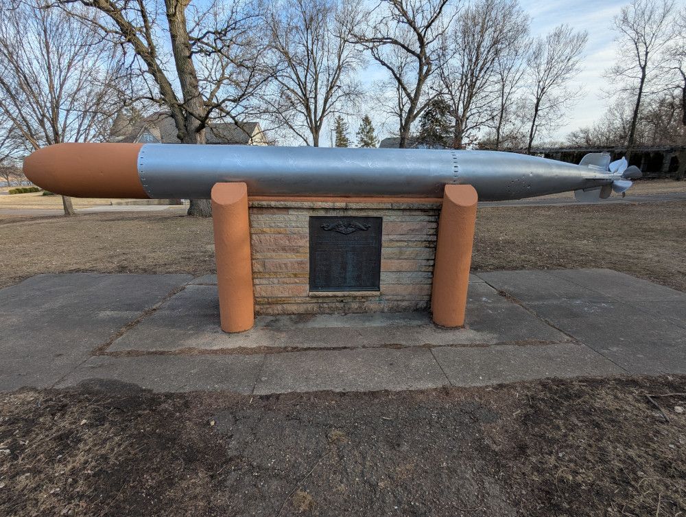 Torpedo mounted on a stand.  A bronze plaque is visible in the center of the stand.