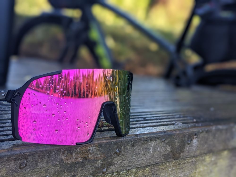 Photo of the reflection on water and tree in pink mirrored sun glasses the sun glasses have rain drops on. The decking (fishing platform) they are resting on is wet and blurred in the background is a bike. 