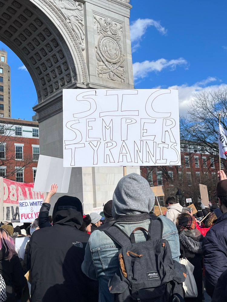 From behind, man holding a sign in Latin Sic Semper Tyrannis. Wash Sq Arch in background. 