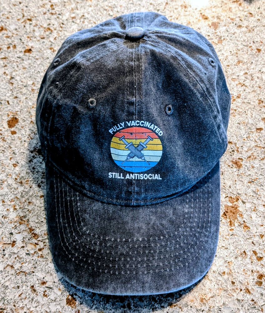 A blue denim hat with an image of two crossed syringes. 
The words written on the hat are: "Fully vaccinated. Still Antisocial."