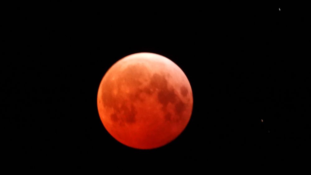 A photo of a full moon during a lunar eclipse. The majority of the moon has a red-orange tone, while a very small area at the top of the moon appears lighter.