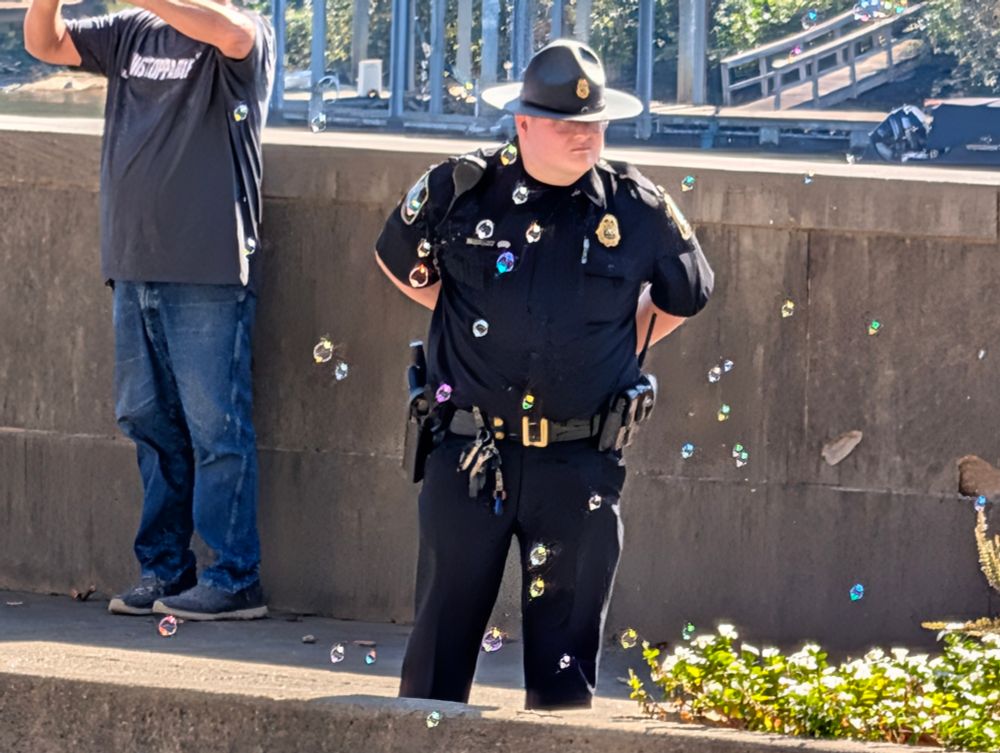 A security guard with his arms behind his back stands on a sidewalk with soap bubbles floating in the foreground.