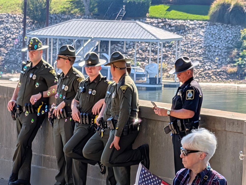Bored police lean against a concrete wall while bubbles float in the foreground. To the right of the police, also bored and leaning against the wall, a security officer looks at his phone. In the lower right foreground a woman wearing sunglasses holds a small American flag.