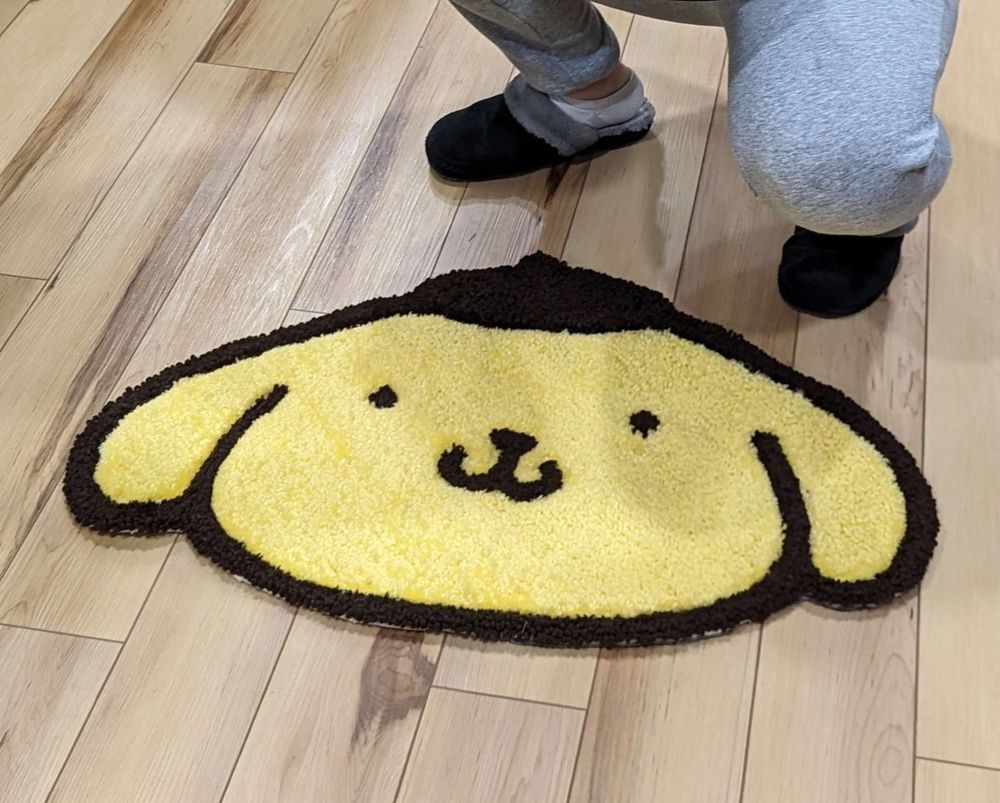 A punch needle rug of Pompompurin sitting on a light-colored hardwoof floor in front of its recipient. The photo is cropped so that we only see a person's lower-half.