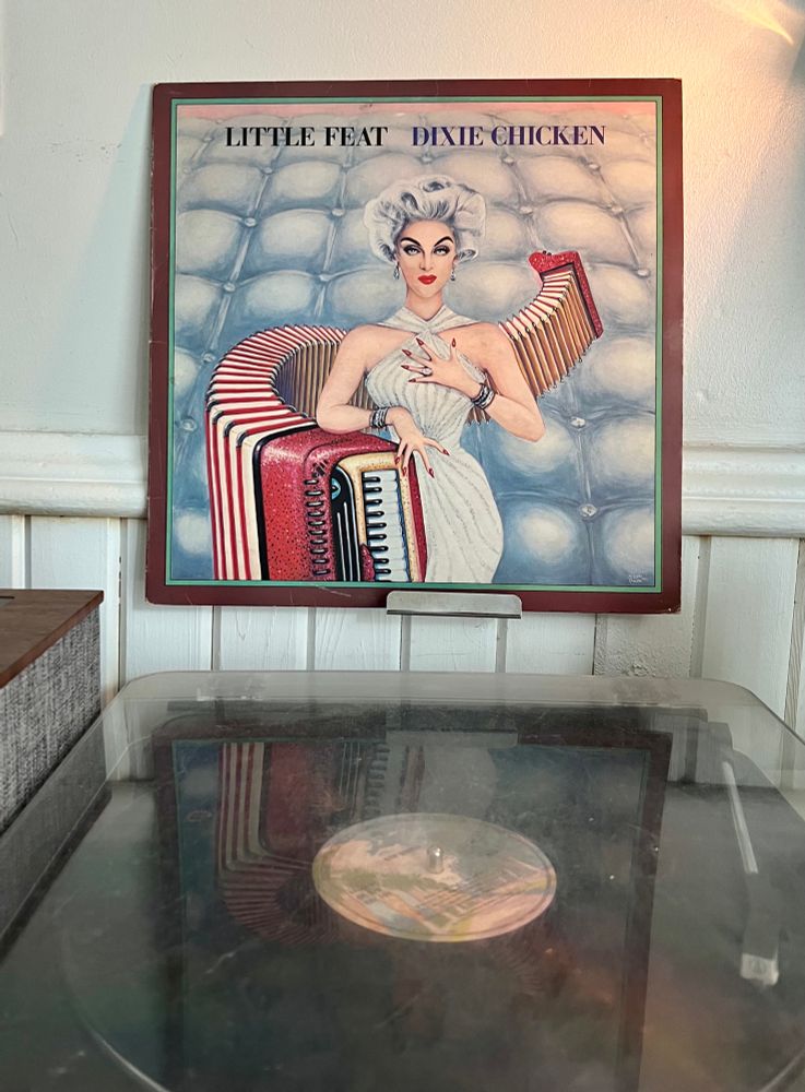 Turntable playing Little Feat’s album Dixie Chicken with the album cover featuring a drawing of a woman in a white dress leaning on an accordion. 