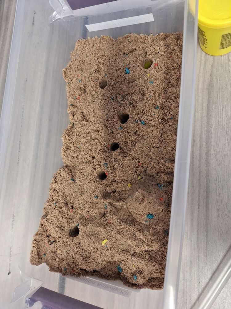 A bin full of sand with 6 uniform small holes cut out.