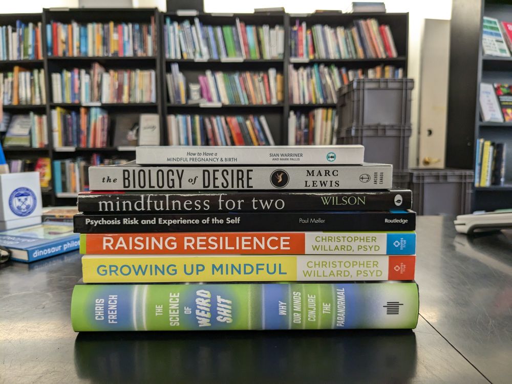 A stack of 7 books on a bookstore checkout counter: How to Have a Mindful Pregnancy and Birth by Sian Warriner, The Biology of Desire by Marc Lewis. Mindfulness for Two by Kelly G. Wilson, Psychosis Risk and Experience of the Self by Paul Moller, Raising Resilience and Growing Up Mindful both by Christopher Willard, and The Science of Weird Shit by Chris French