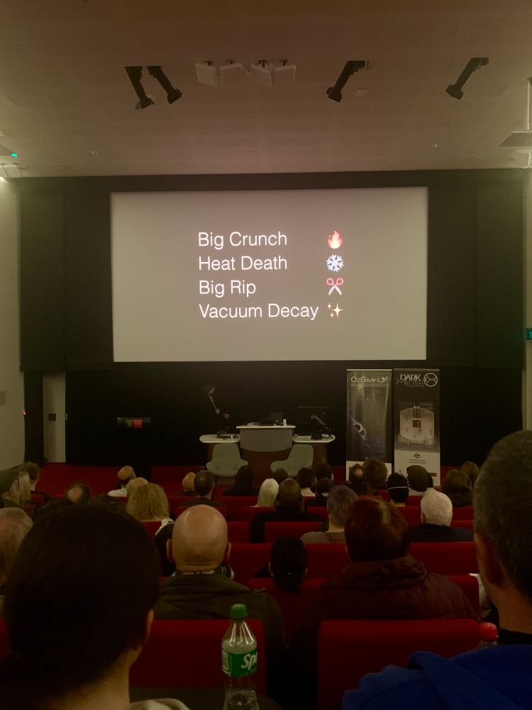 Image of a darkened lecture hall with a projected slide highlighting some of the ways our Universe could meet its end. The darkness of the room definitely matched the vibe of this lecture. There are four theories highlighted: Big Crunch, Heat Death, Big Rip or Vacuum Decay. The last one mine and Dr Katie Mack’ favourite! 