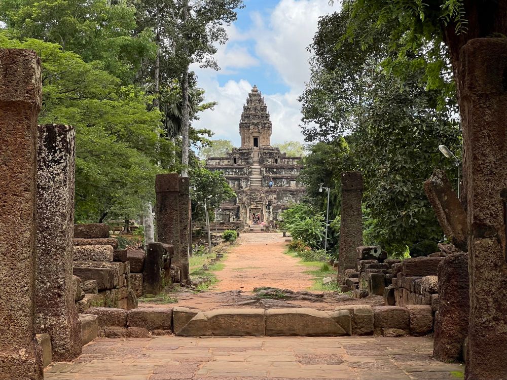 A 5 tiered pyramid style temple with a tower on top, located at the end of a stone then dirt path fringed with tall trees
