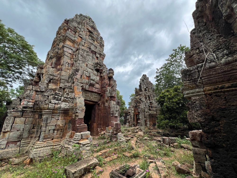 Two crumbling stone temples about 10 meters high, roughly pyramidal with a thick stone framed doorway and dark interior. Ground littered with stone and overgrown with flowering plants
