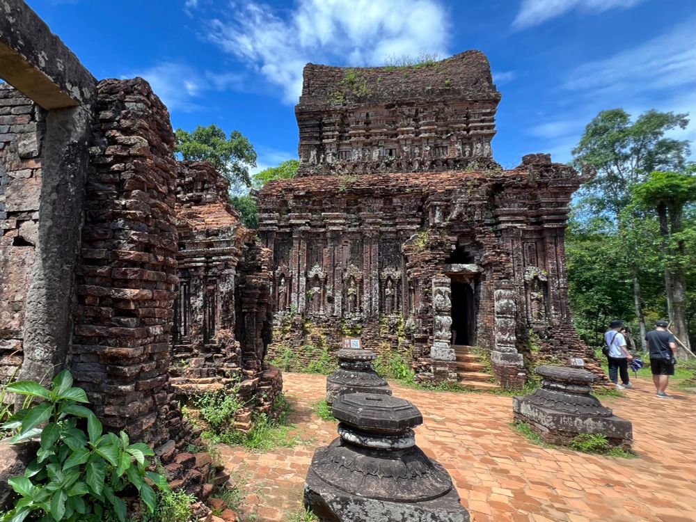 A crumbling brick temple with statues and decorated pillars on either side of a dark doorway. Trees in background